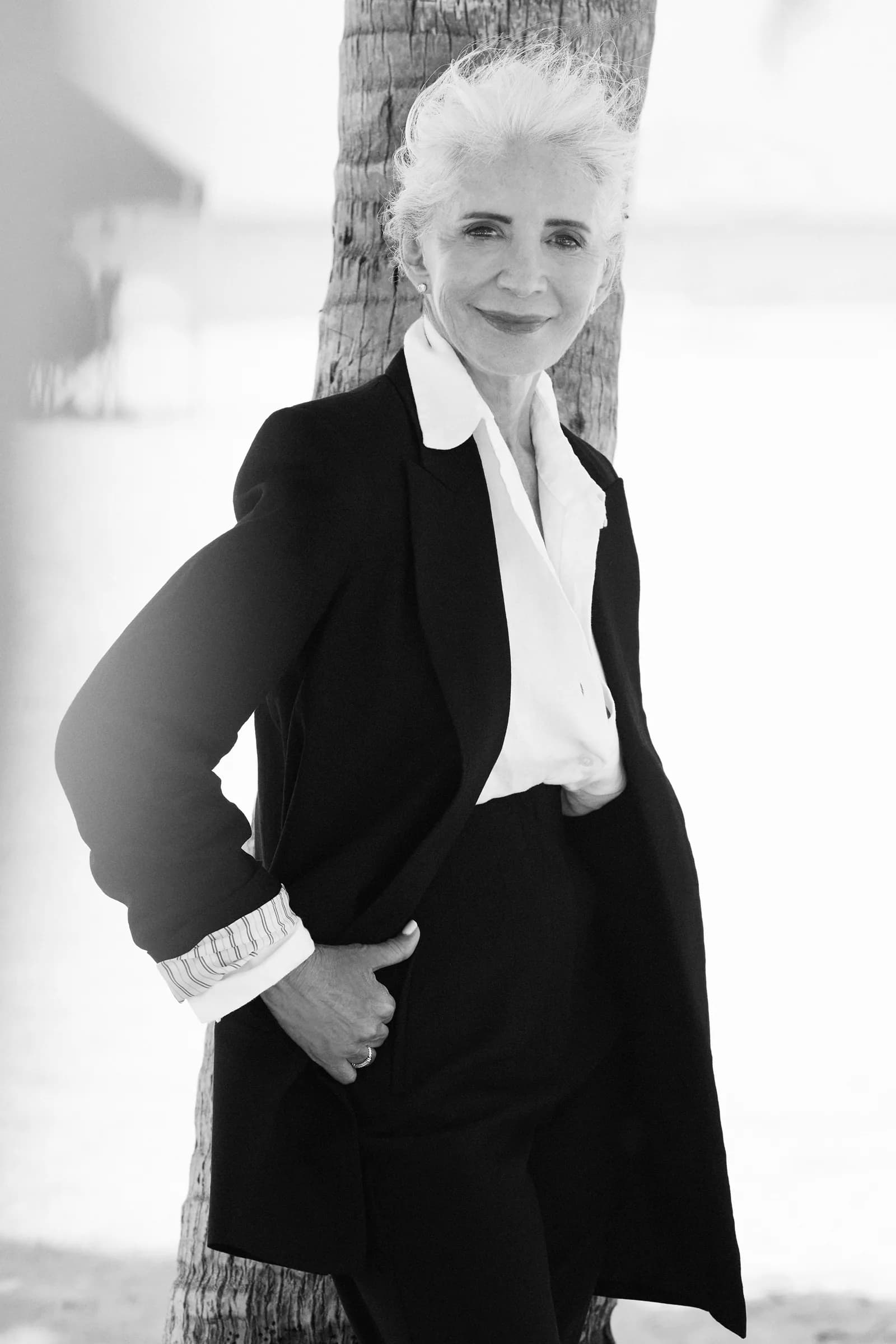 Ruth, founder of Go SILVR Goddess, in a black blazer and white shirt, leaning on a palm tree by the beach